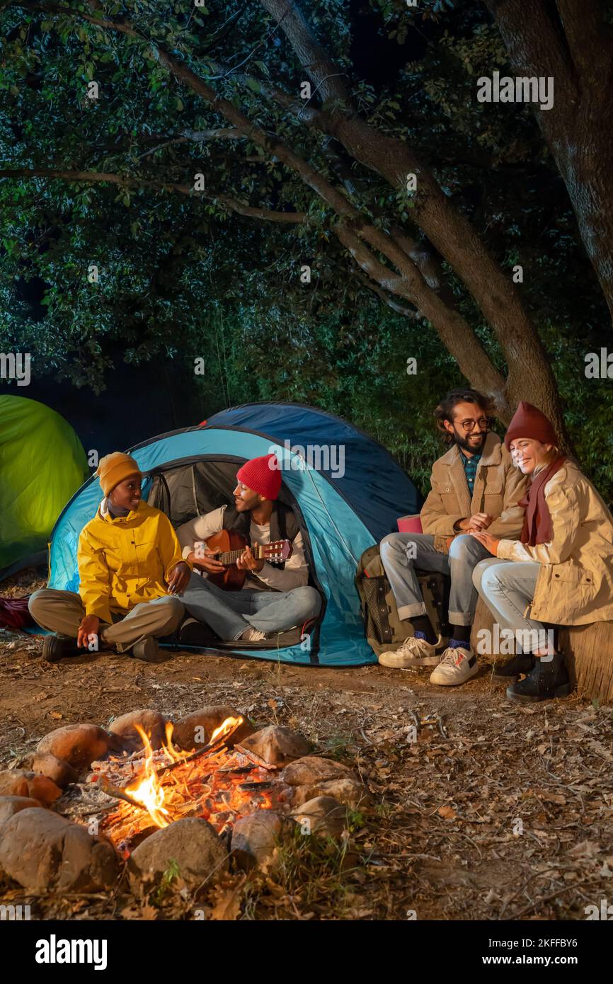 Happy friends having fun together enjoying bonfire camping playing guitar in nature at night. Stock Photo