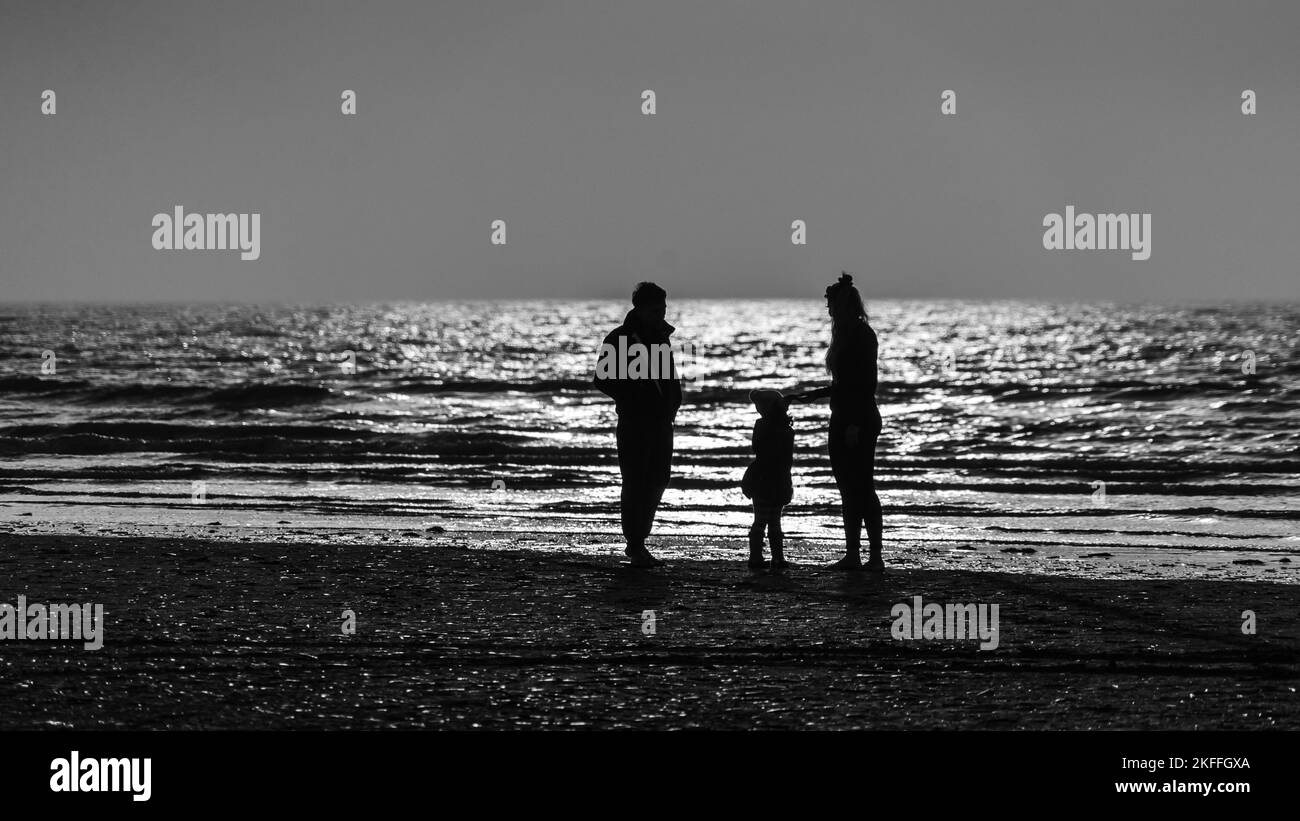 Black and white photograph of a father, mother and their small child standing on the water's edge on a beach before the backdrop of the glittering sea Stock Photo