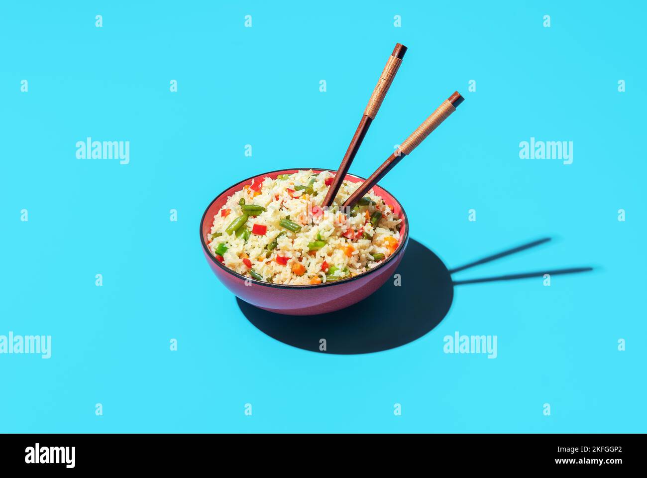 Fried rice with vegetables in a red bowl with chopsticks minimalist on a blue table. Delicious asian dish, fried white rice with a mix of vegetables. Stock Photo
