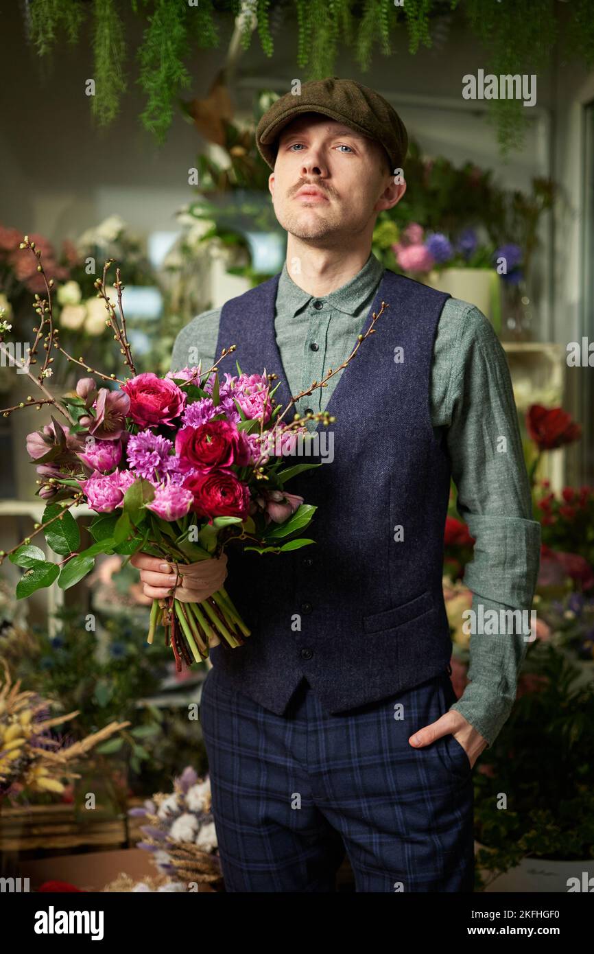 Attractive cheerful caucasian male in peaked cap and vintage clothes from 20s holding beautiful bouquet of red and violet flowers. Mother's Day or Valentine's Day concept. High quality image Stock Photo