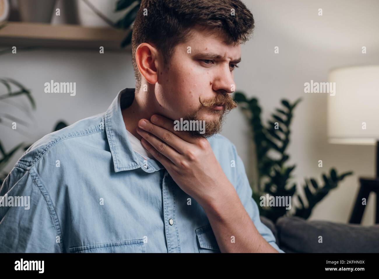 Sick young caucasian man sitting on couch at home touch neck suffer