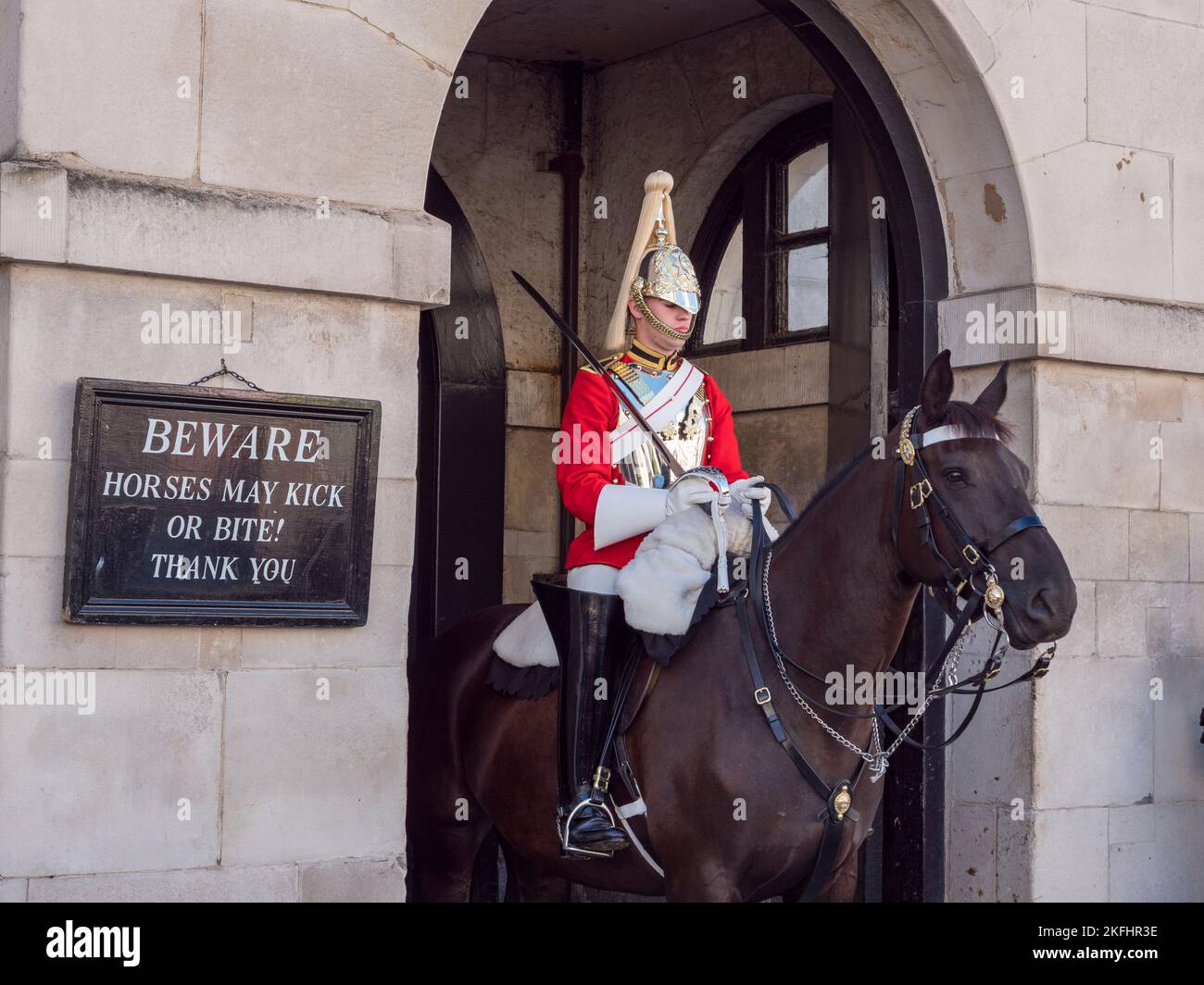 'Beware. Horses may kick or bite' sign beside a mounted Life Guard on duty at the Horse Guards barracks on Whitehall, London, UK. Stock Photo