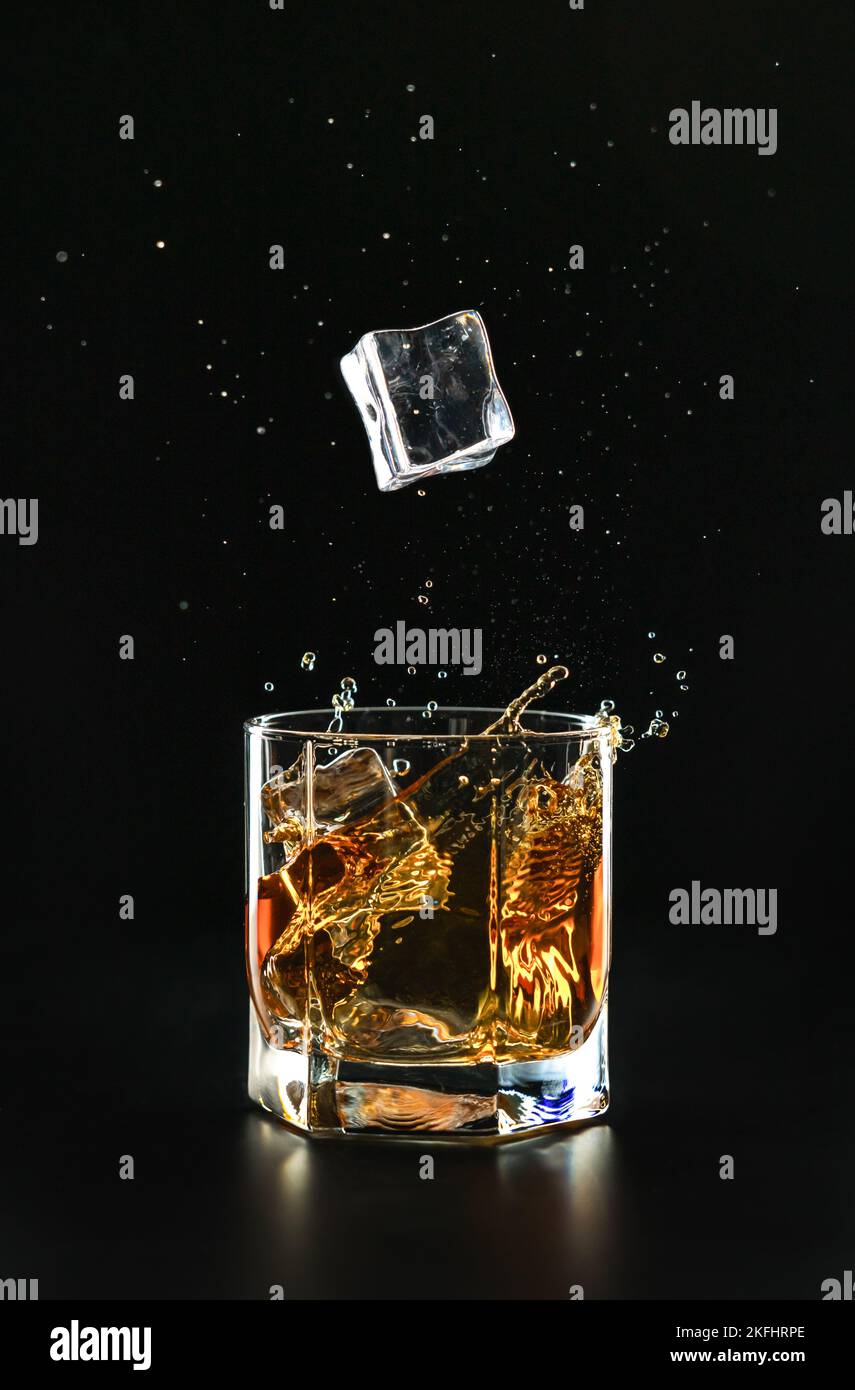 An ice cube falls into a glass of poured whiskey causing a splash and ...