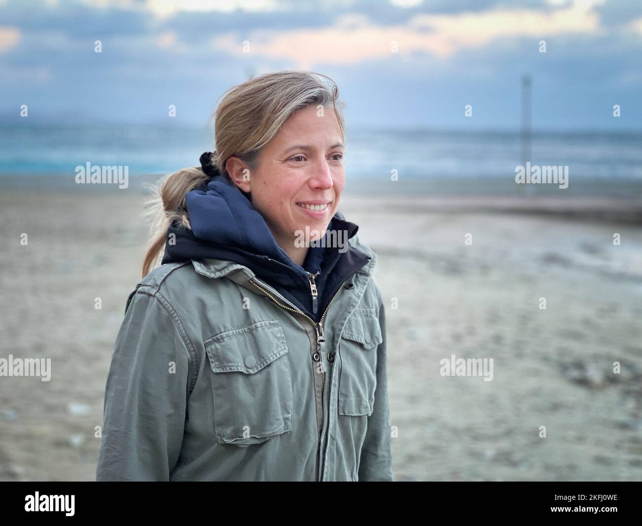 Smiling Caucasian mid adult woman wearing winter coat looking away while standing at beach against cloudy sky Stock Photo