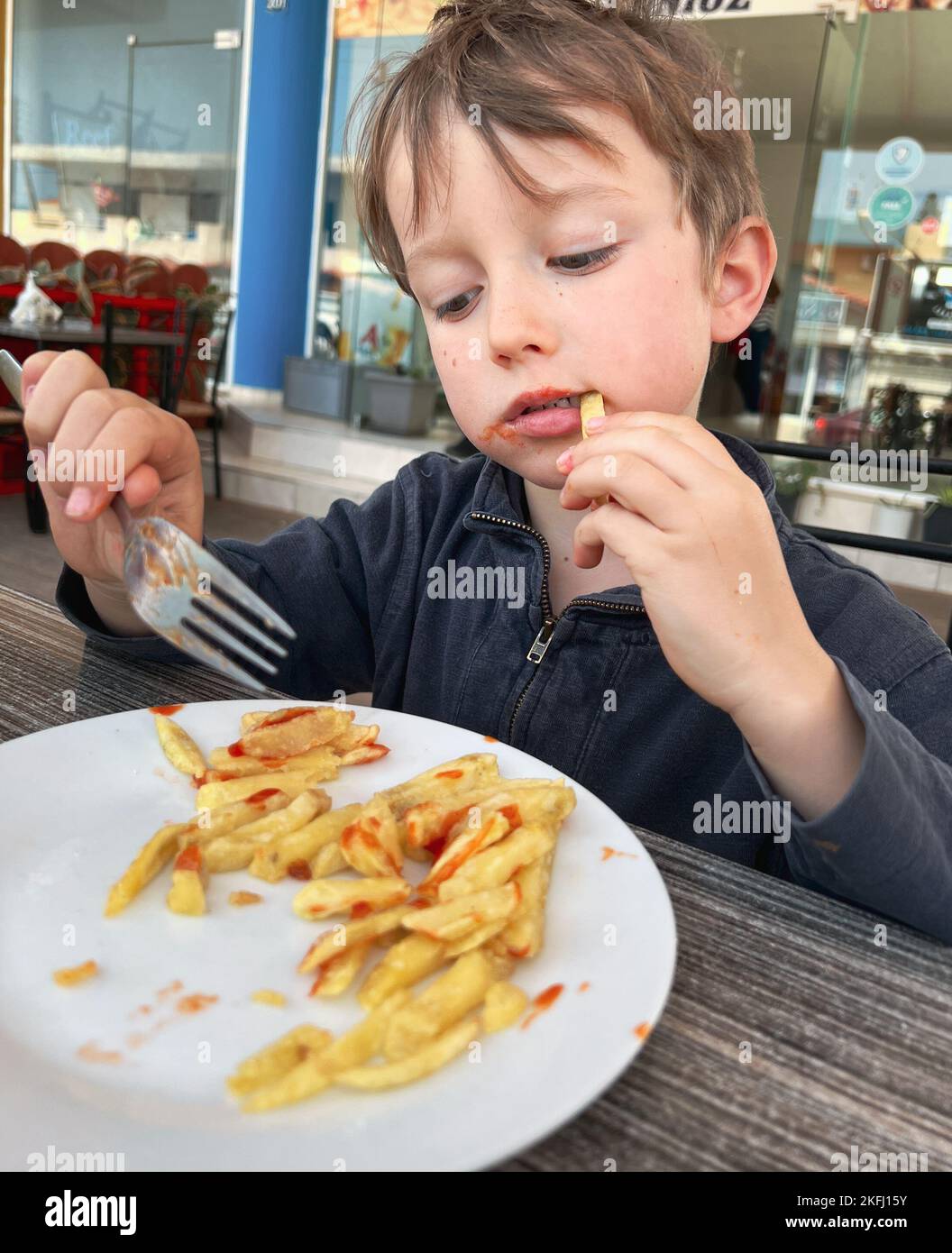 Caucasian cute boy holding fork and eating french fries with hand at table in fast food restaurant Stock Photo