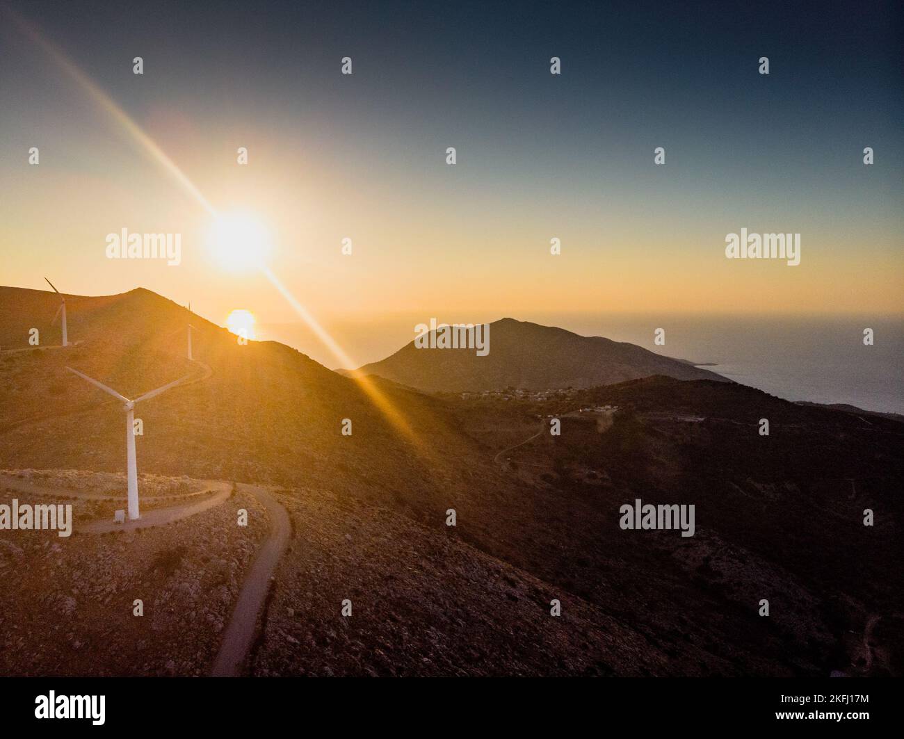 Wind turbines on mountain against sky and scenic view of silhouette ranges against bright sun during sunset Stock Photo