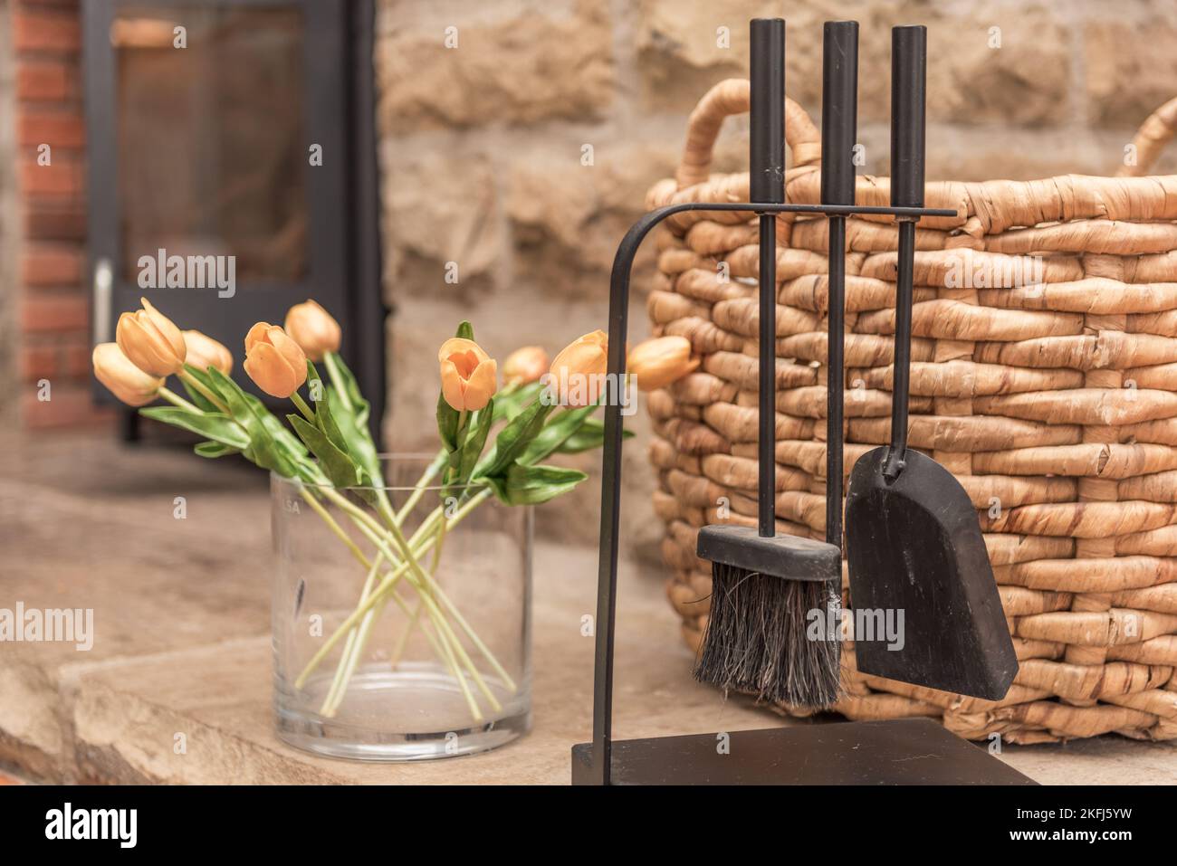 Stone fire hearth with black metal coal brush and shovel. Wicker coal basket. Shallow depth of field with fire stove in the background. Stock Photo