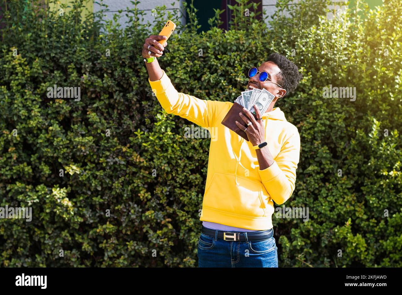 A smiling young African man wearing a yellow sweatshirt, blue jeans and sunglasses is taking a selfie with his mobile phone while holding a wallet wit Stock Photo