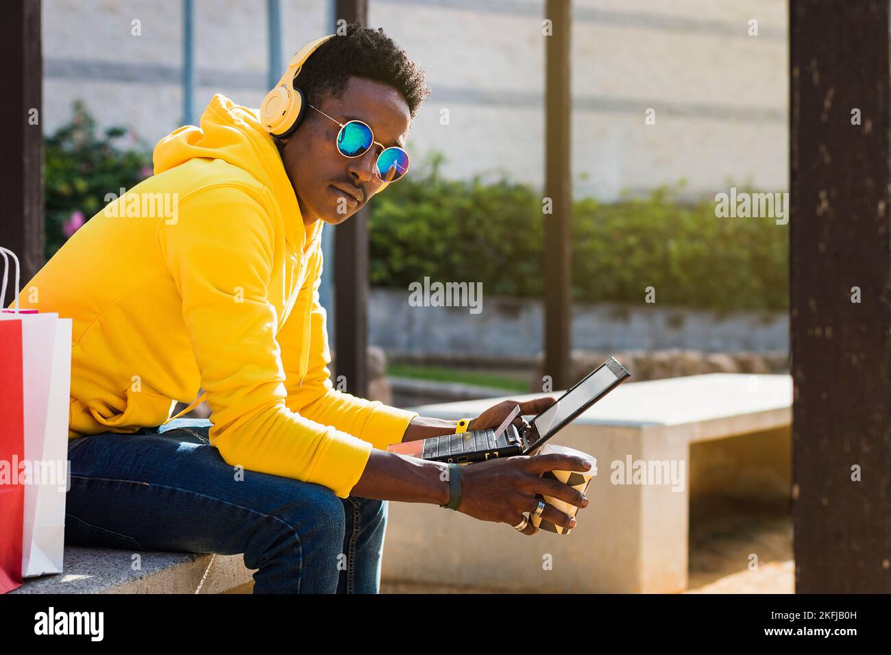 A young African man dressed in a yellow sweatshirt, blue jeans and sunglasses sits on a park bench holding a laptop and a glass of coffee. Above the k Stock Photo
