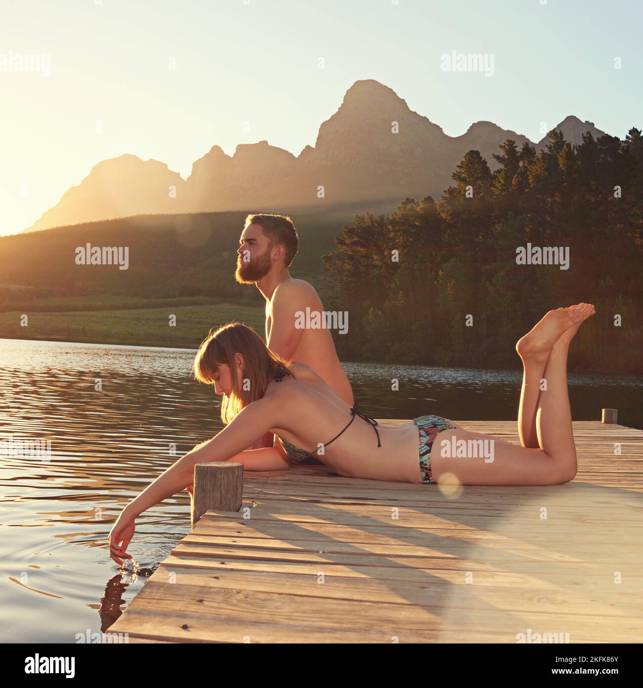 The water is perfect...an affectionate young couple in swimsuits sitting on a dock at sunset. Stock Photo