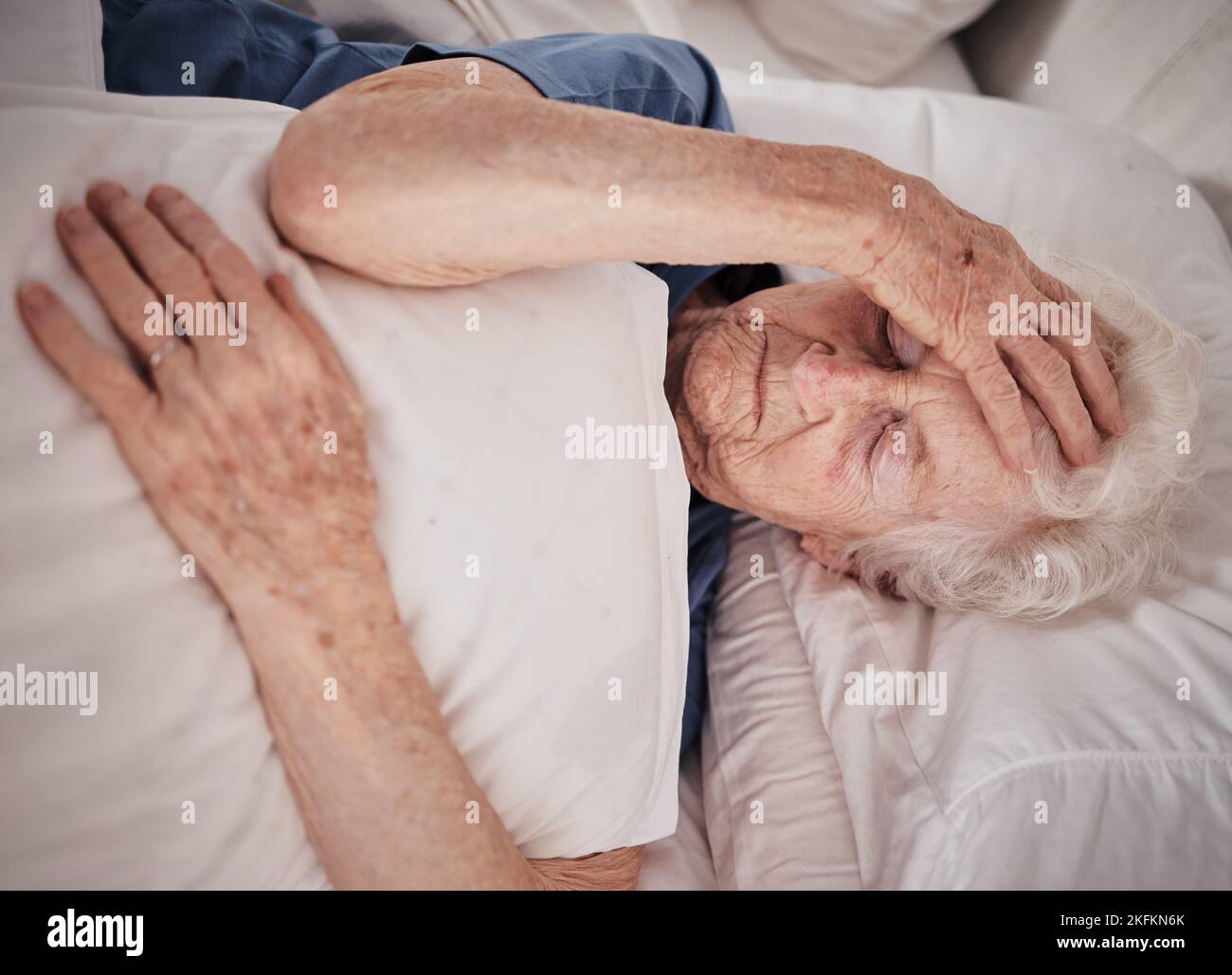 Headache, pain and senior woman in bed for trauma recovery, rehabilitation or rest in elderly care nursing home. Healthcare problem, medical emergency Stock Photo