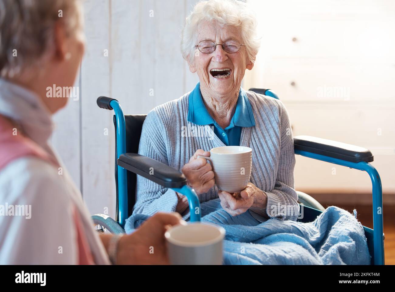 Senior woman, friend and coffee or tea while in a wheelchair for a disability or rehabilitation and happy or laughing about funny conversation or joke Stock Photo