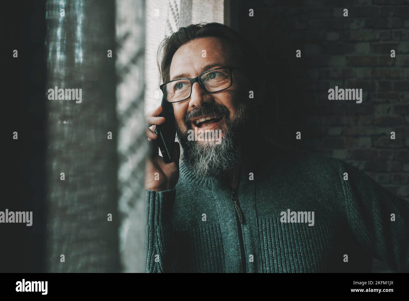 One happy young mature man speaking at the phone smiling and having fun at home near the window. Concept of people and mobile cell communication. Male Stock Photo