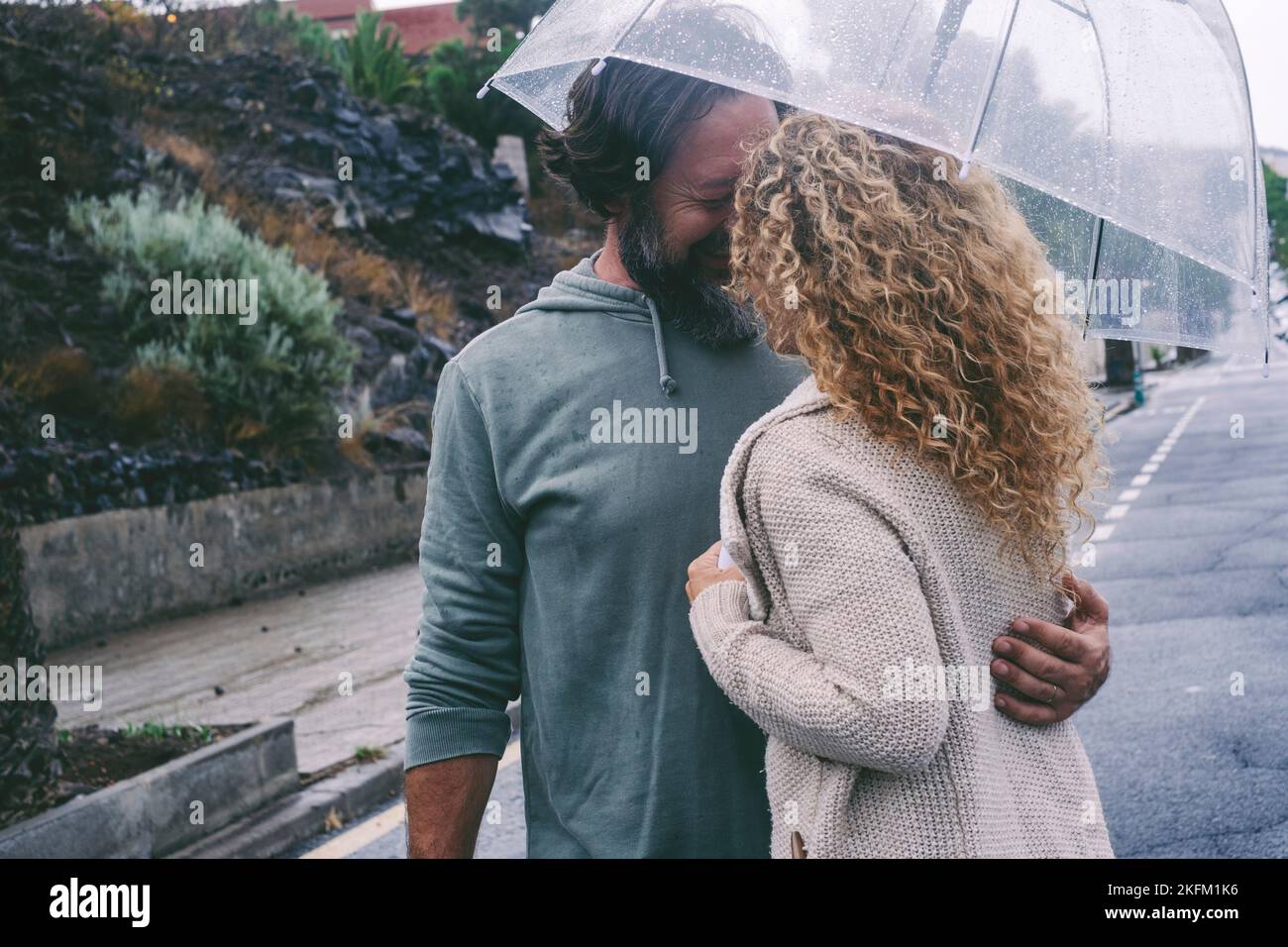 Couple enjoy outdoor leisure activity in rainy day of bad weather hugging and kissing under a transparent umbrella with road street urban in backgroun Stock Photo