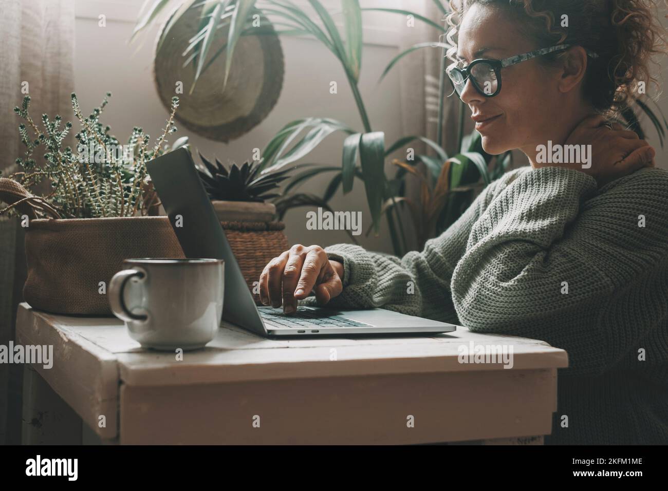 Happy and serene woman with eyeglasses surf the web on a computer at home. Green mood color. Plants in background. Adult female people working on lapt Stock Photo