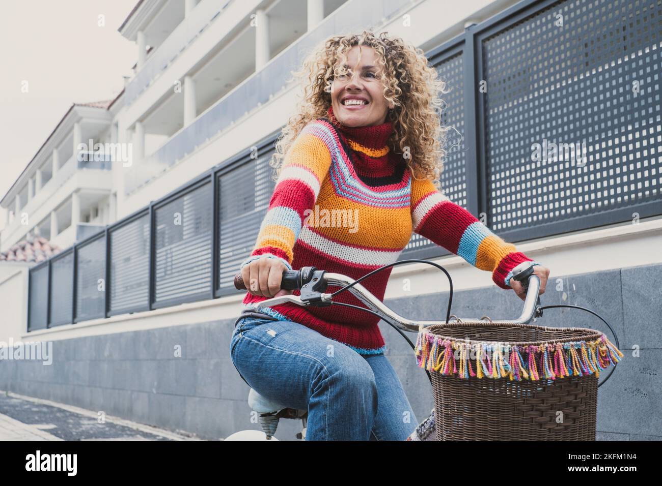Happy middle age active woman riding a bike in the city wearing colorful sweater. Green transport mode and outdoor urban leisure activity with pretty Stock Photo