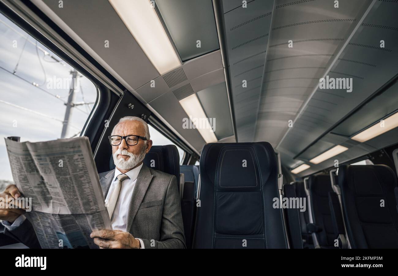 Senior entrepreneur reading newspaper while sitting by the window. Male professional is traveling in modern passenger train. He is wearing suit while Stock Photo