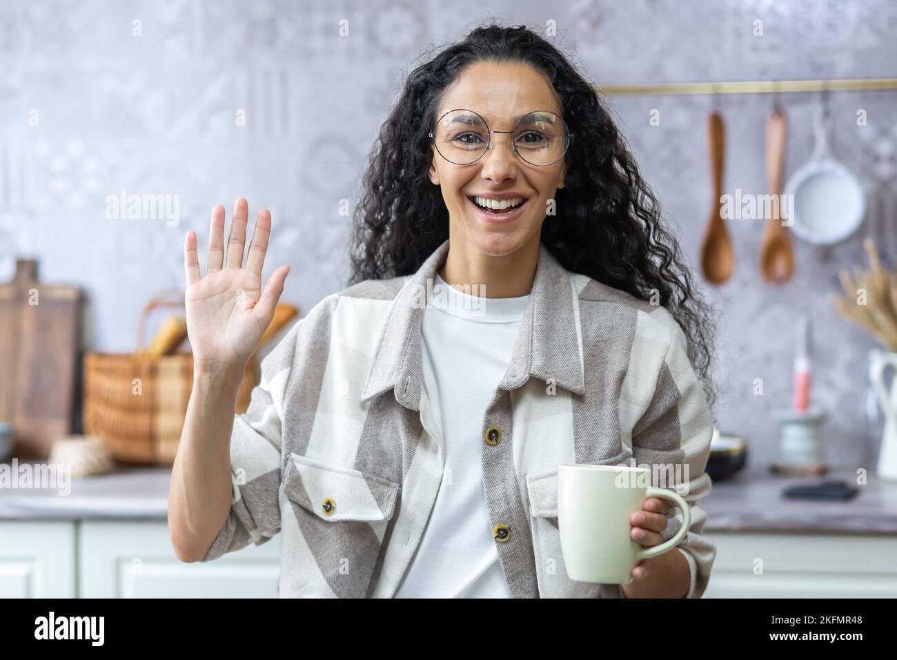 Young beautiful hispanic woman talking and waving at the camera during a video call. Standing in the kitchen at home, holding a cup, smiling, runs his blog. Stock Photo