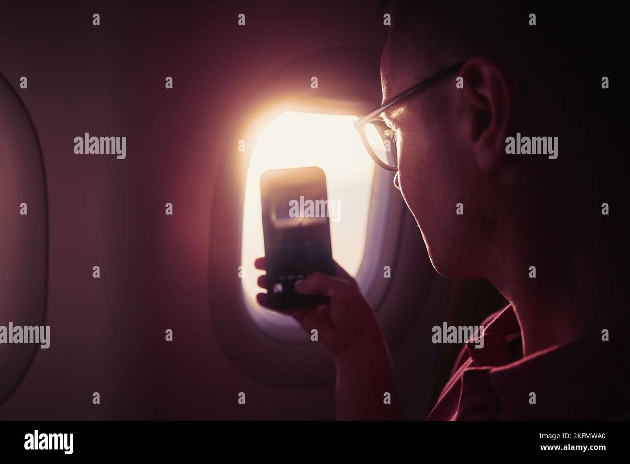 Portrait of man while taking pictures through airplane window. Passenger using phone during flight at beautiful sunset. Stock Photo