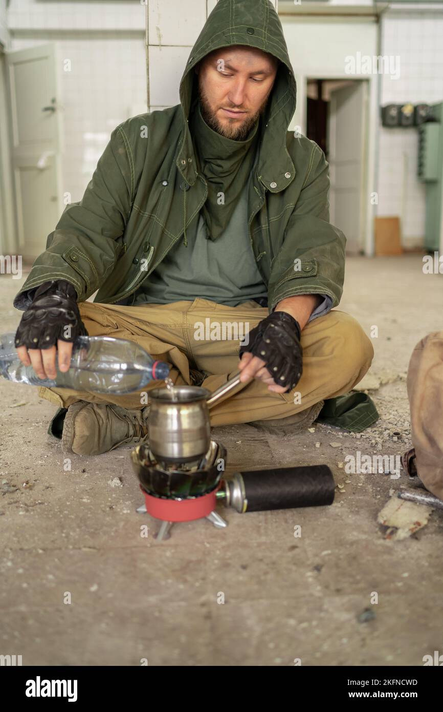 A survivor in a post-apocalyptic world. A man in a ruined building sits on the floor, pours and heats water in a mug to keep warm. Cold, alone, traged Stock Photo