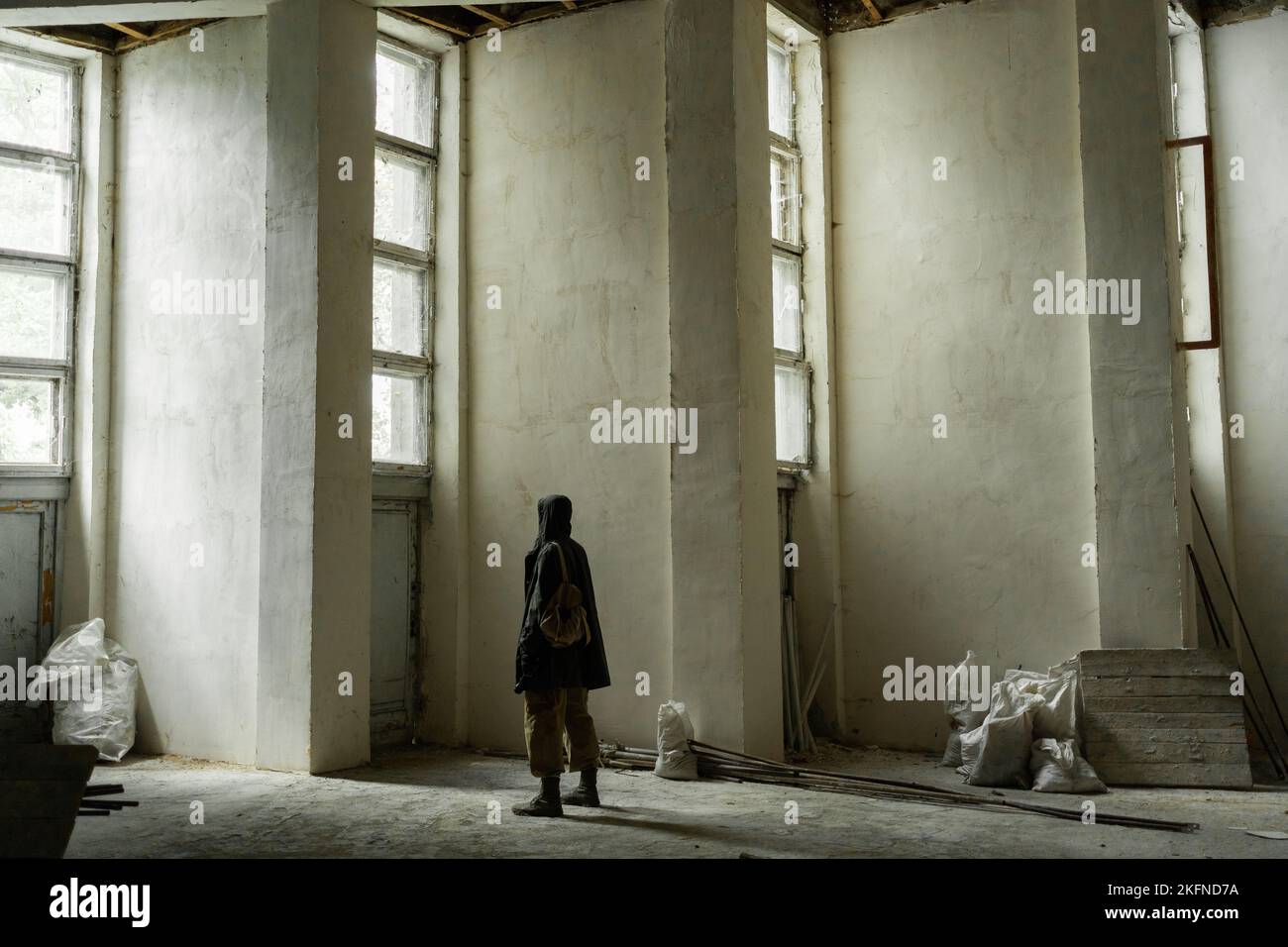 In an old abandoned building stands a lone traveler with a hood on his head, without a face. the concept of a survivor after the third world war in se Stock Photo
