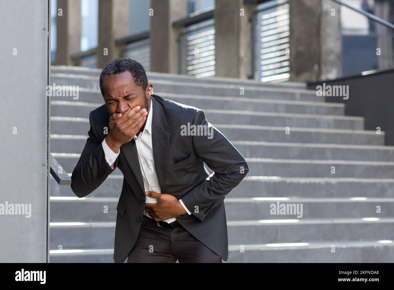 Nausea and pain in the stomach, a businessman from outside the office building is sick, a man is holding his hand on his stomach in a business suit, he got sick with poor-quality food. Stock Photo