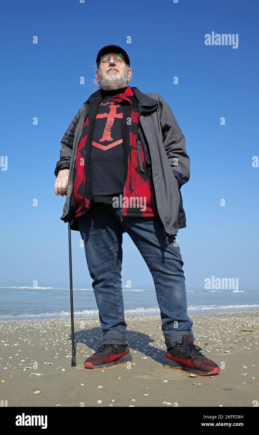 Senior man standing on the beach. He wears a shirt with a templars cross. It's beautiful sunny day in spring Stock Photo