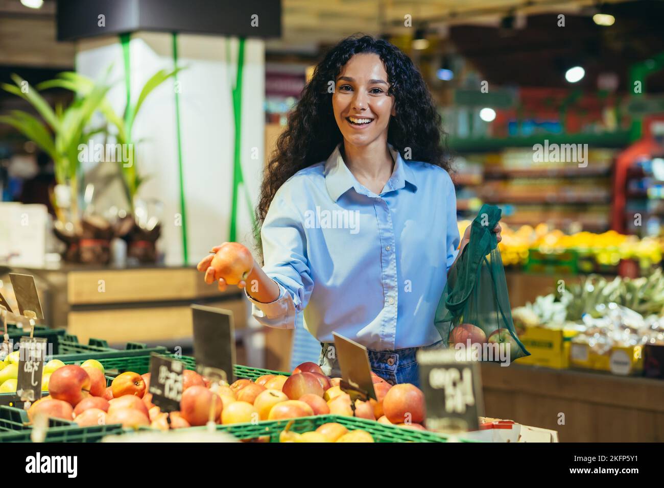 Portrait of happy woman shopper in supermarket, Hispanic woman chooses apples and fruits smiling and looking at camera, with grocery basket chooses goods. Stock Photo