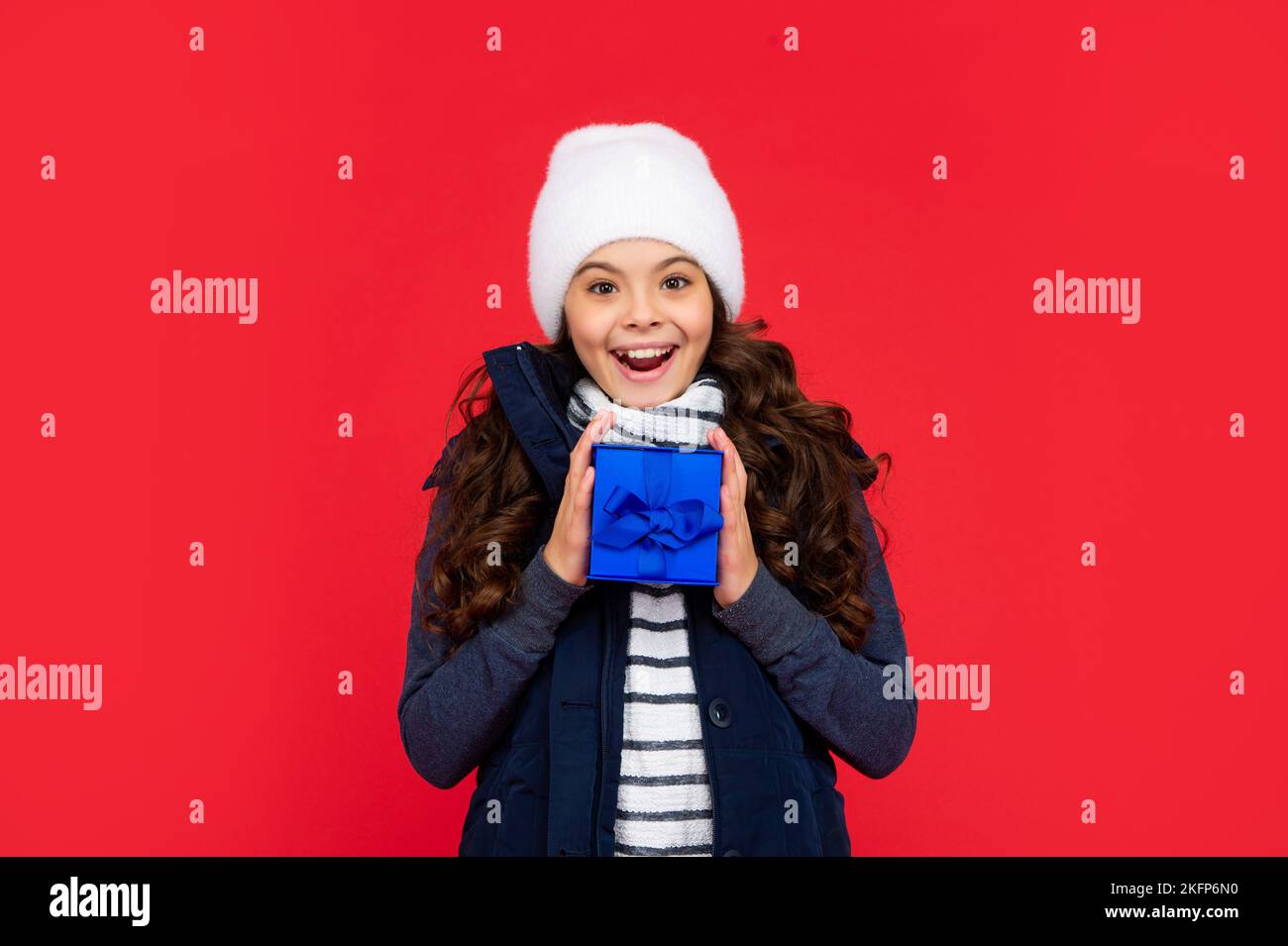 amazed child in puffer jacket and hat hold box. kid with present. teen girl on red background. Stock Photo
