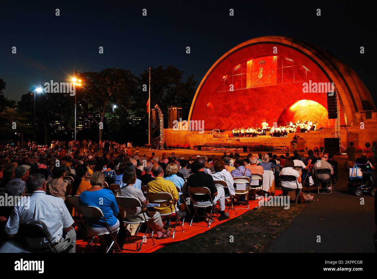 A  audience enjoys a concert by a symphony orchestra playing classical music at night in the illuminated Hatch Shell in Boston Stock Photo