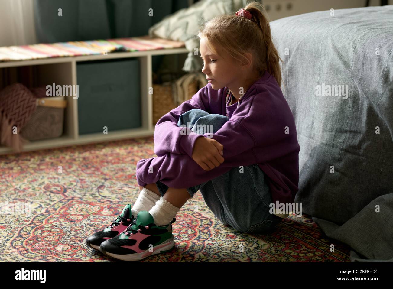 Little girl with sad expression sitting on floor along in her room Stock Photo
