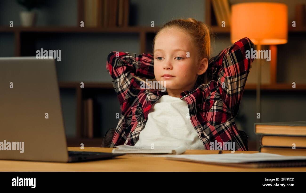 Little preschooler pupil girl watch tutor lesson by video call e-learning take break during homework education put hands behind head feel relaxed Stock Photo