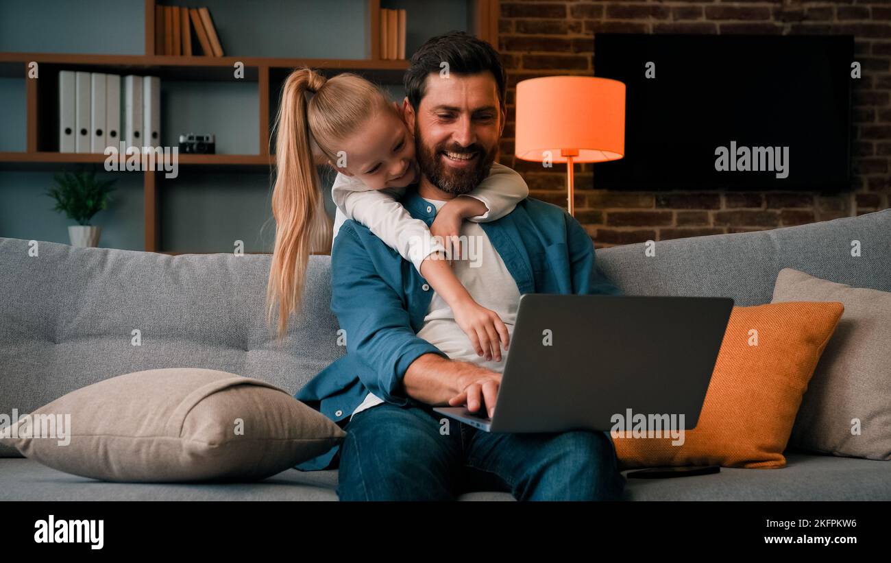 Cute active naughty girl noisy child plays with daddy hugs father distract parental attention disturbs parent from work on laptop at home single Stock Photo