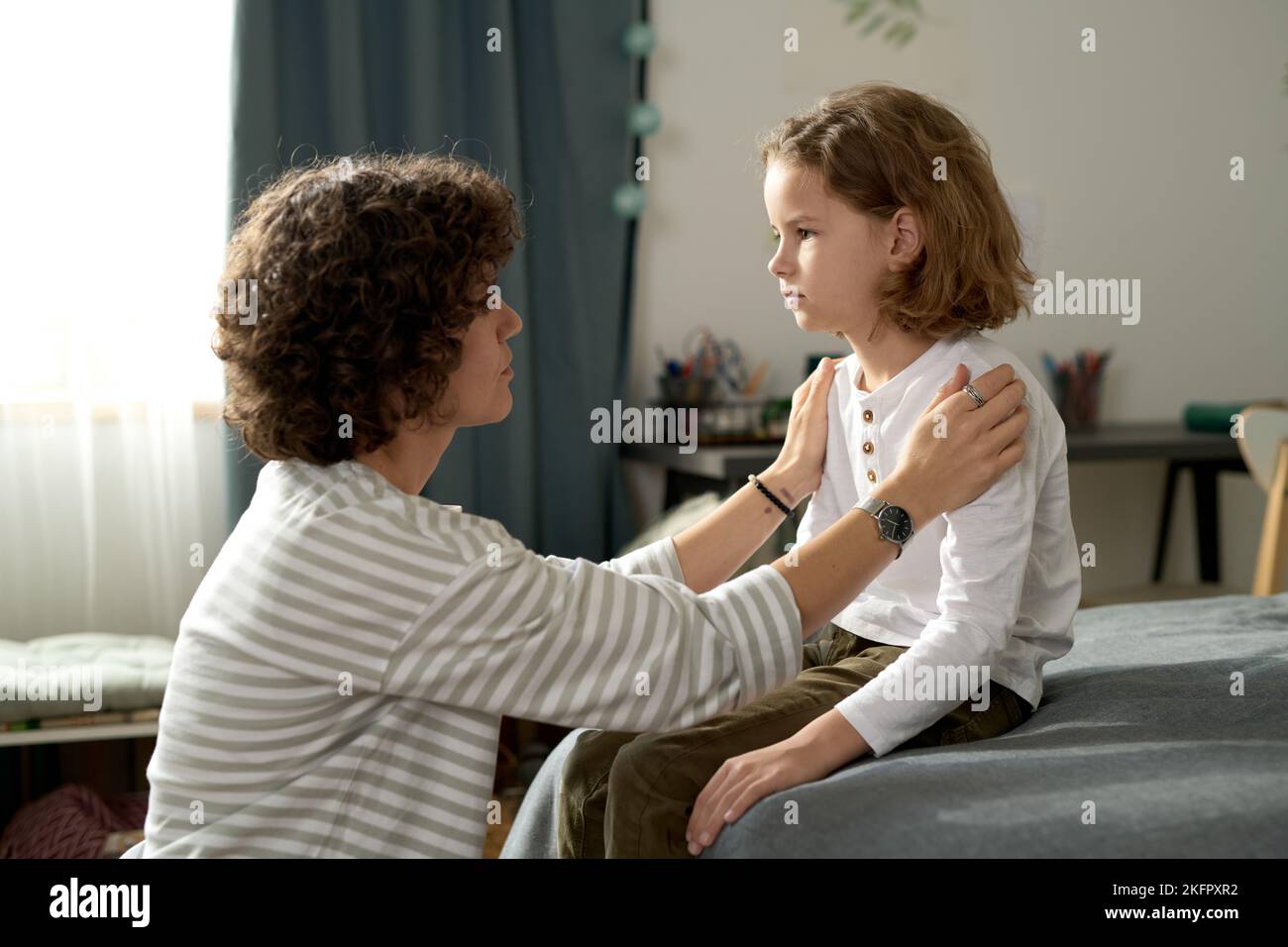 Mother sitting in front of her child and talking to him about his behavior while he sitting on bed Stock Photo