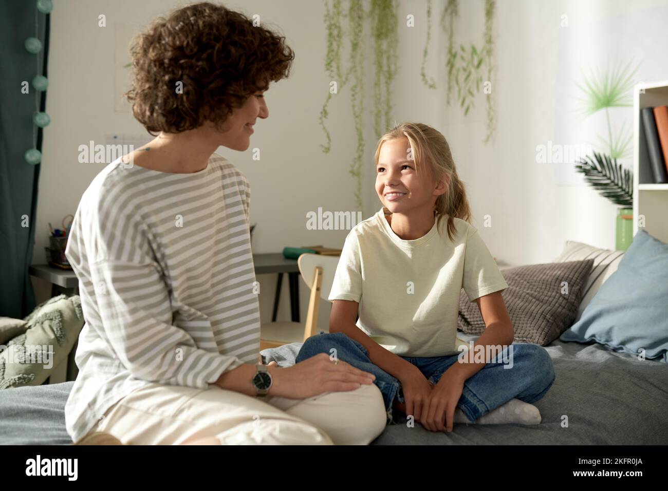 Mother talking to her little daughter about female secrets, they sitting on bed in bedroom and smiling Stock Photo