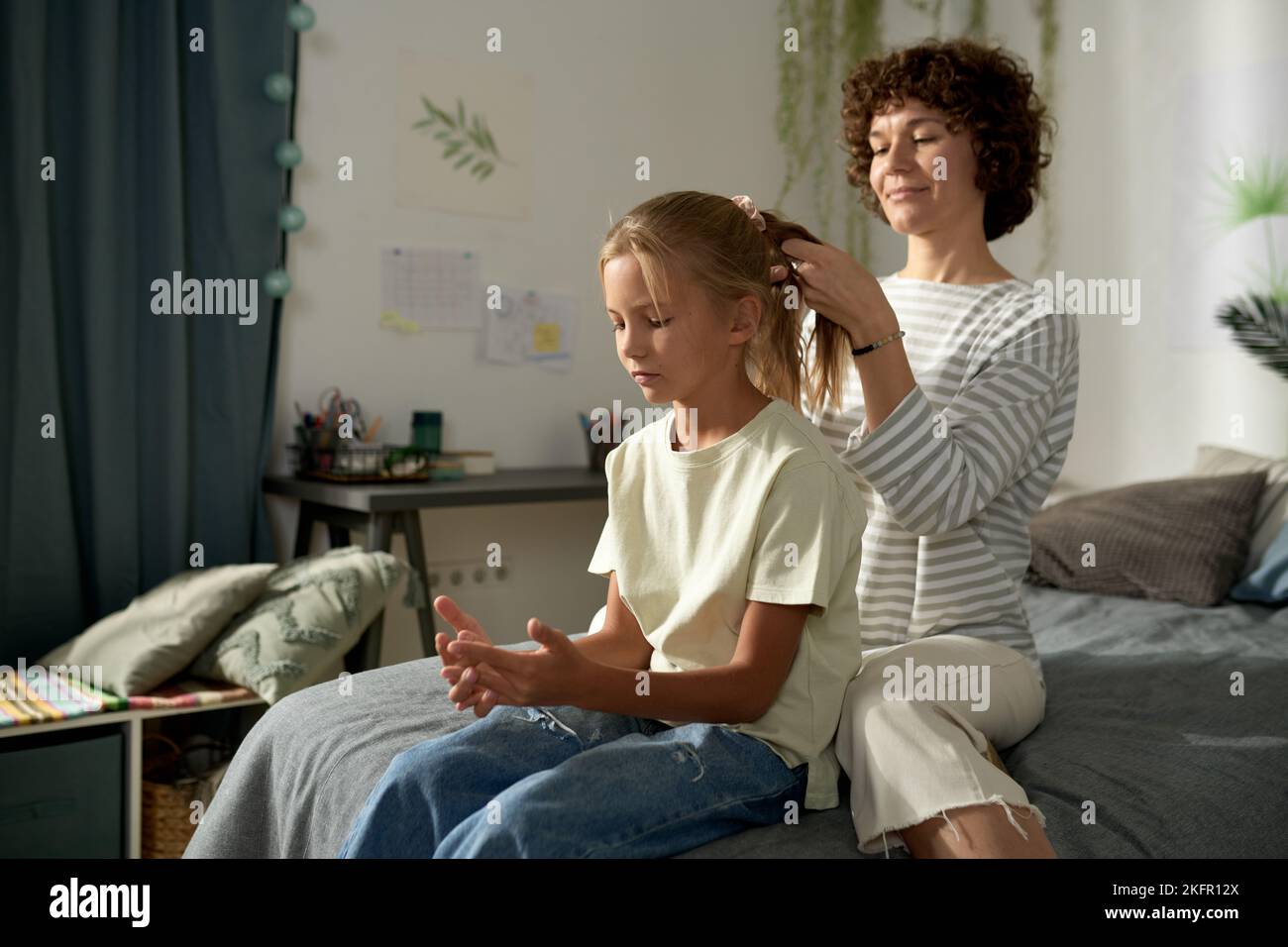 Young mother making hairstyle to her daughter in bedroom, they spending leisure time together Stock Photo