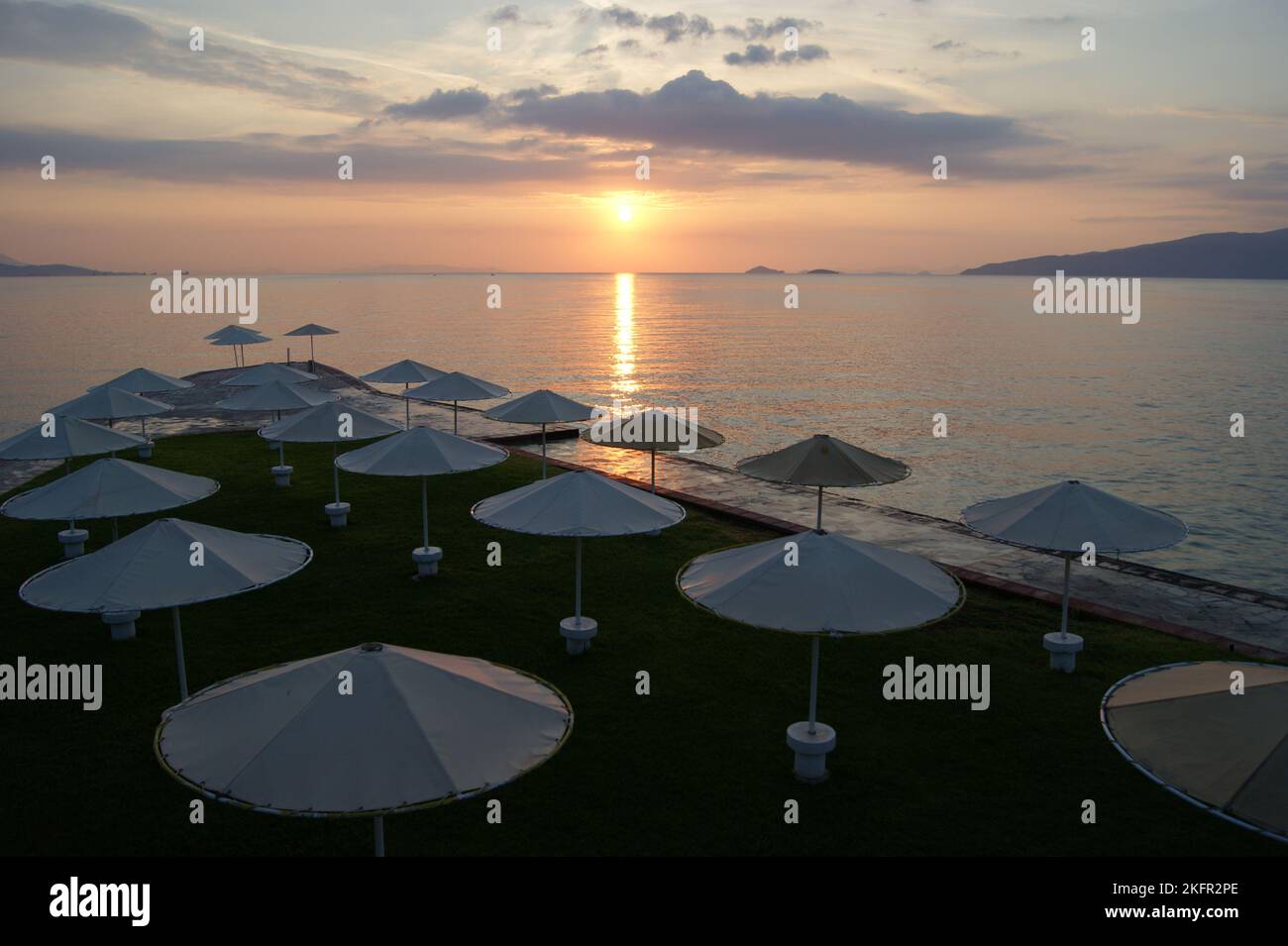 Beach umbrellas, view of the sun setting in the Aegean sea, orange sky background, at Kalamaki Beach, Saronic Gulf of the Aegean, Isthmia, Greece Stock Photo