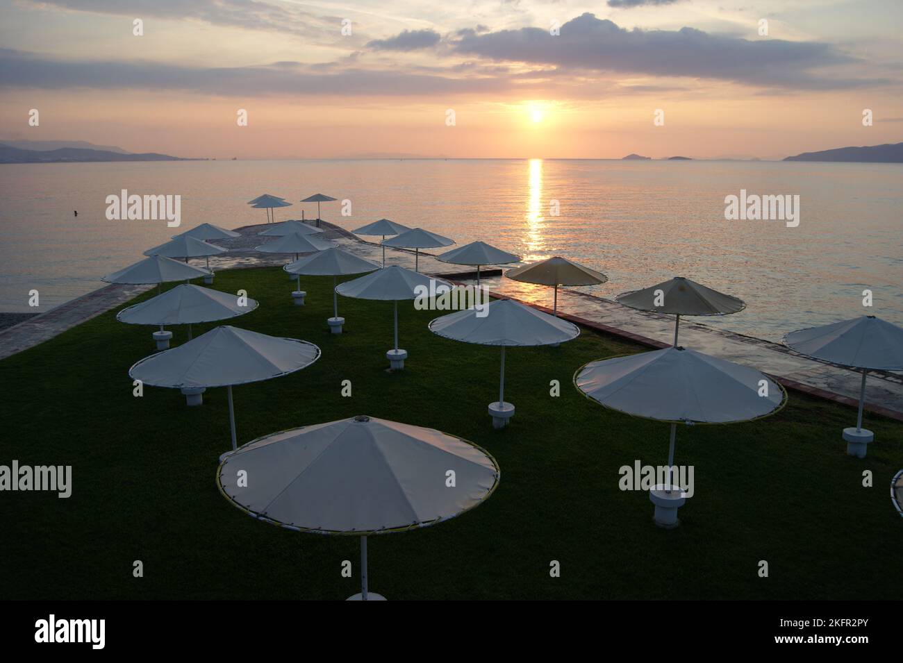 Beach umbrellas, view of the sun setting in the Aegean sea, orange sky background, at Kalamaki Beach, Saronic Gulf of the Aegean, Isthmia, Greece Stock Photo