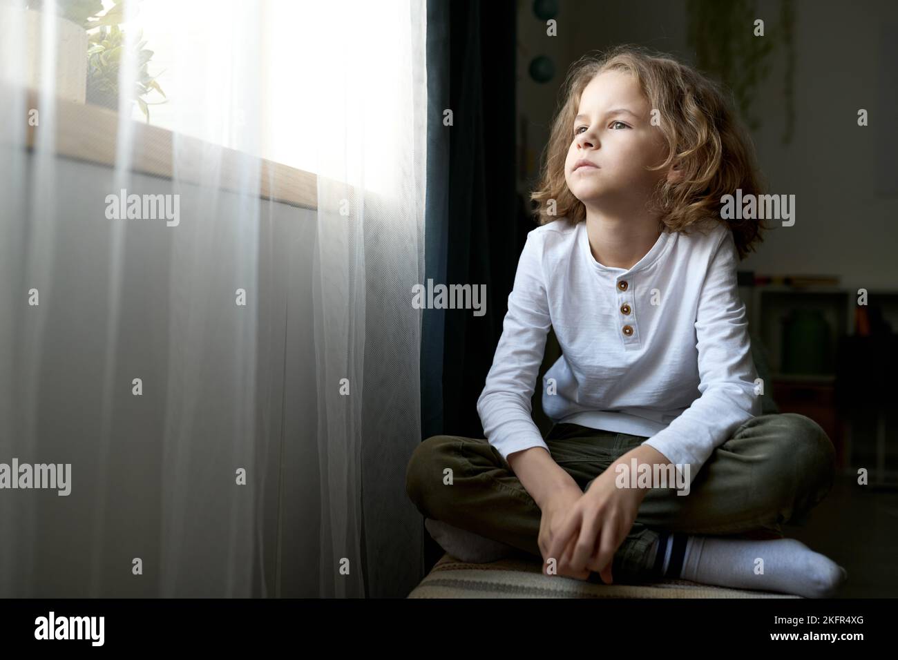 Pensive little boy sitting in his room and looking at window with thoughtful sight Stock Photo