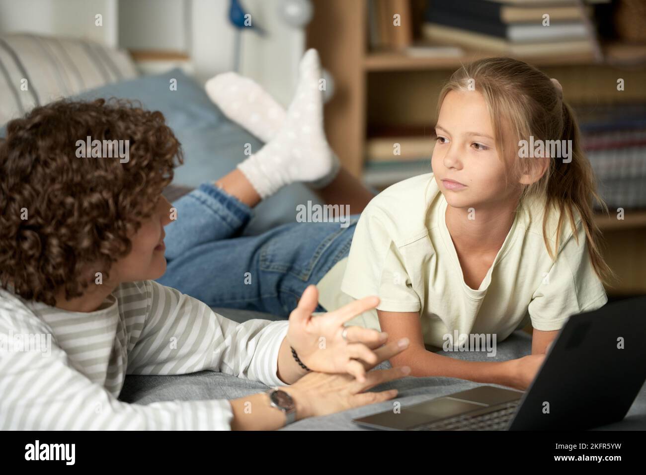 Little girl listening to her mom while she explaining her something, they lying on bed and using laptop Stock Photo