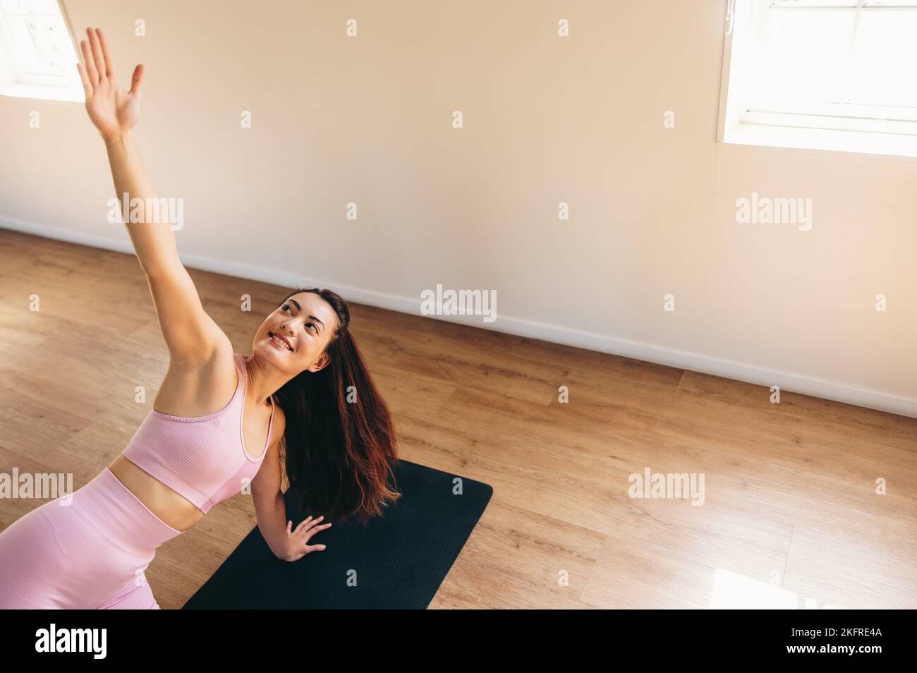 Happy young woman stretching her arm while practicing hatha yoga in a fitness studio. Sporty young woman smiling while doing the side angle plank on a Stock Photo