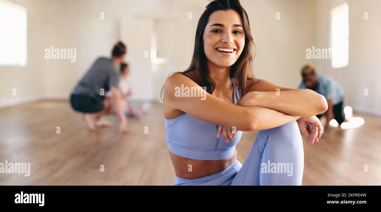 Portrait of a fit young woman smiling at the camera while sitting in a yoga studio with her class in the background. Happy young woman having a workou Stock Photo