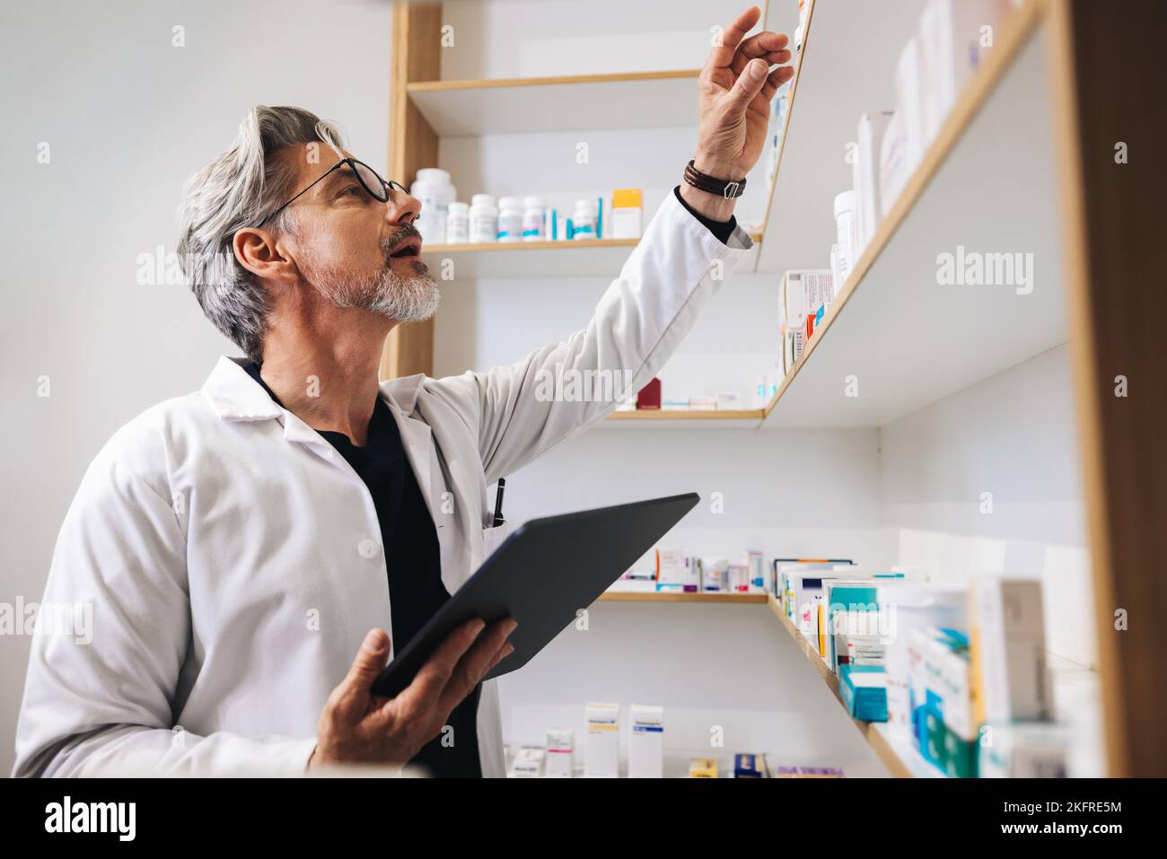 Senior pharmacist getting prescription medication from a shelf in a chemist. Male healthcare worker using a digital tablet to fill online prescription Stock Photo