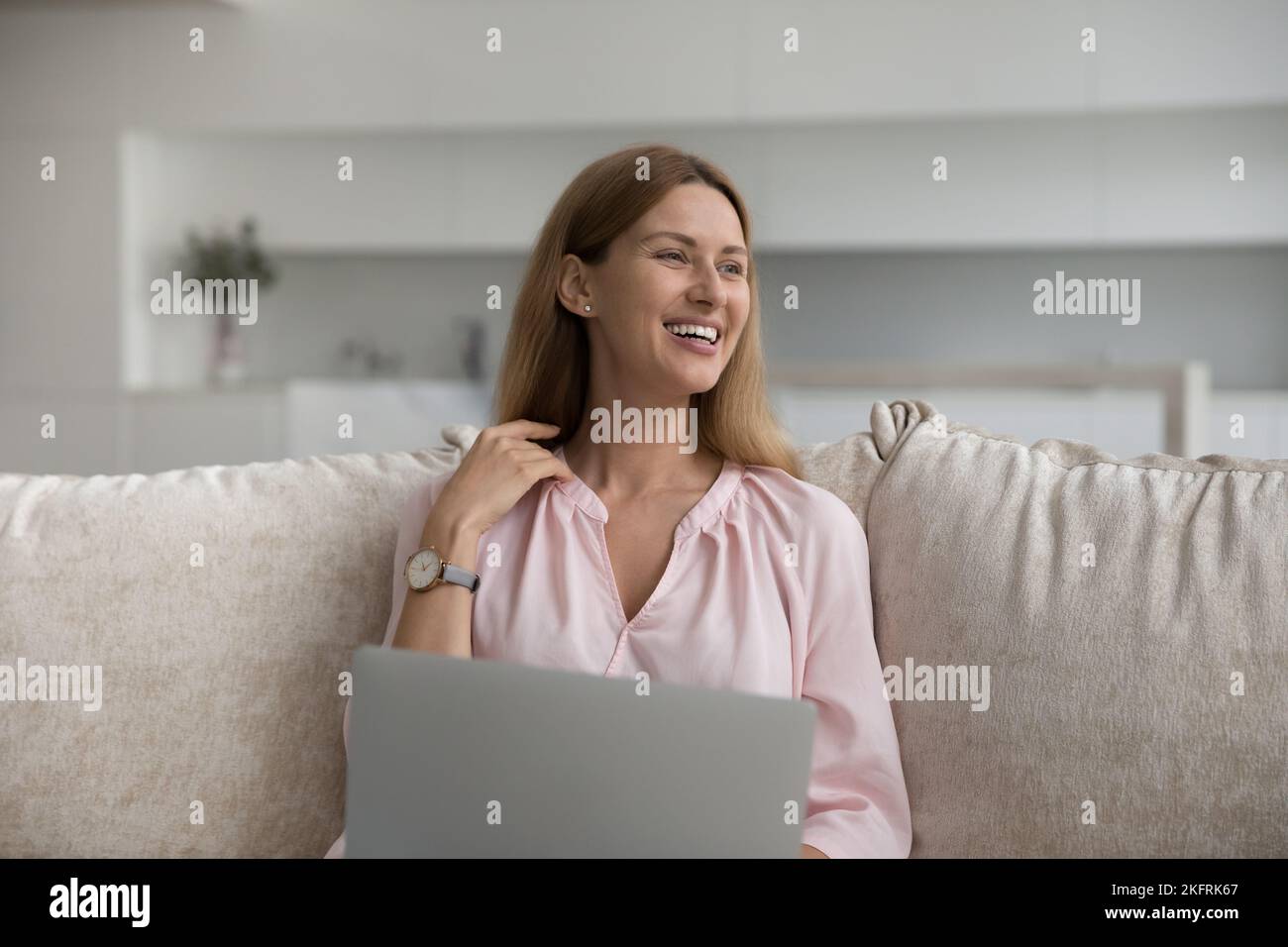 Smiling young woman sitting on couch at home using laptop Stock Photo