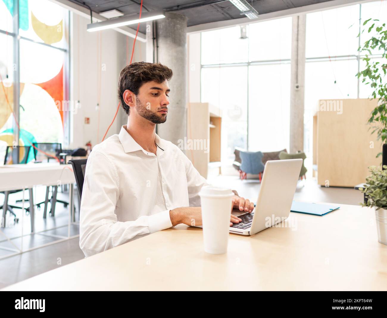 Concentrated young bearded man in white shirt sitting at table and browsing internet via laptop while working on laptop in spacious office cafeteria Stock Photo