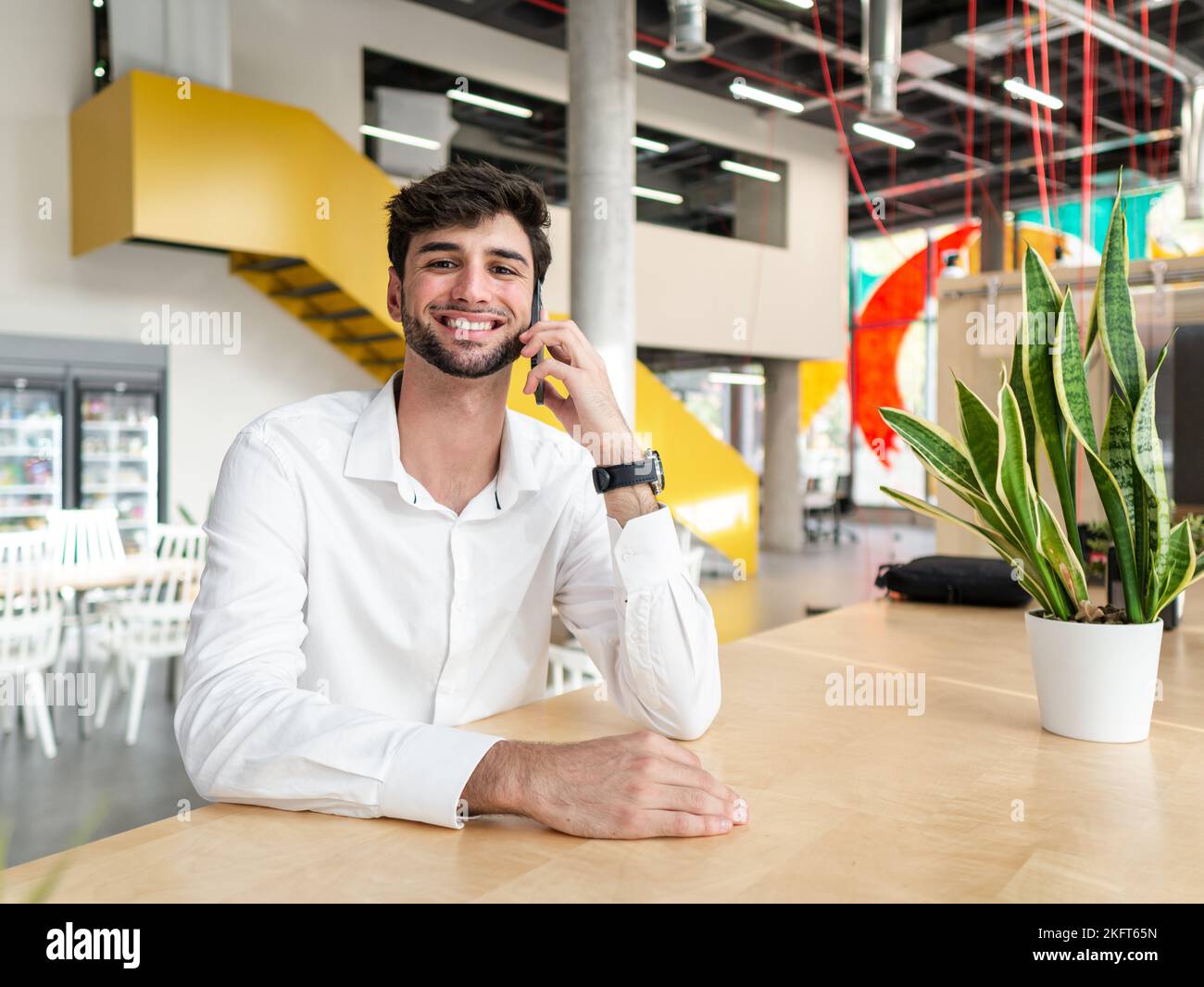 Smiling young bearded man in smart casual outfit standing near table and talking on smartphone while looking at camera in modern workspace Stock Photo