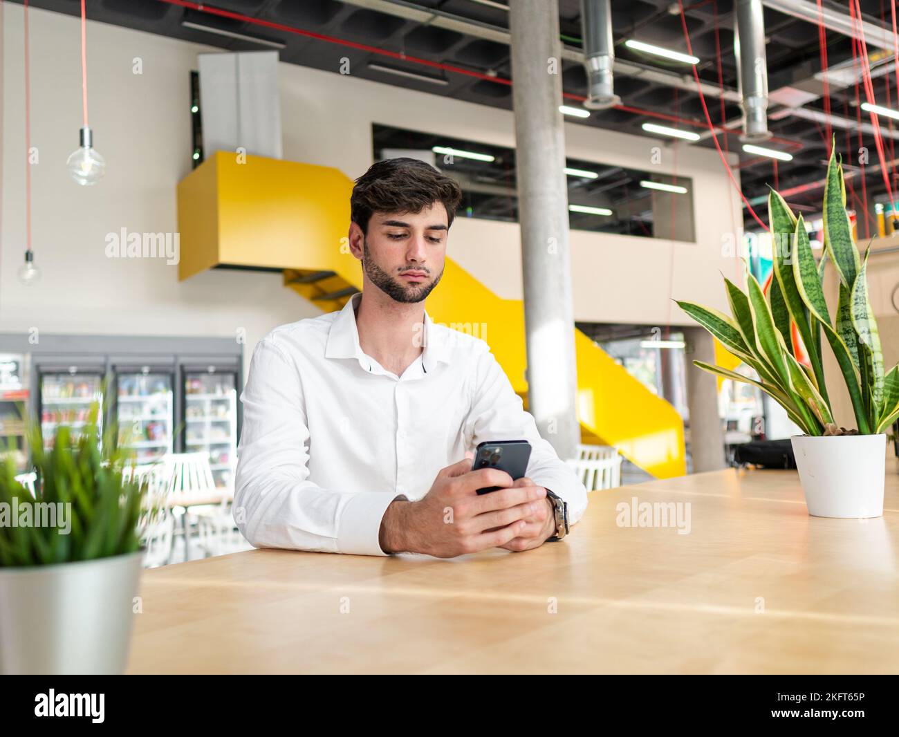 Concentrated young bearded man in white shirt sitting at table and texting message via cellphone in light modern workspace Stock Photo