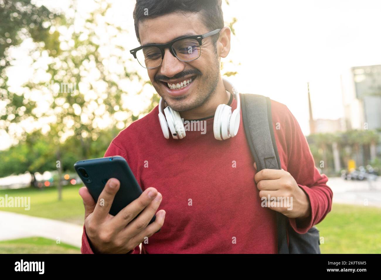 Smiling young man in red sweatshirt and headphones on neck with rucksack standing on city street browsing on mobile phone Stock Photo