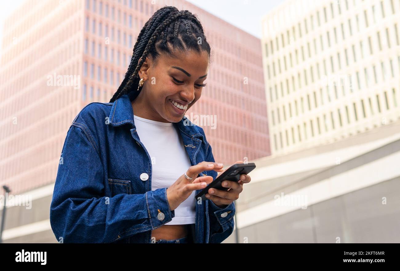 Low angle of African American female browsing mobile phone messaging on social media while standing against modern building Stock Photo