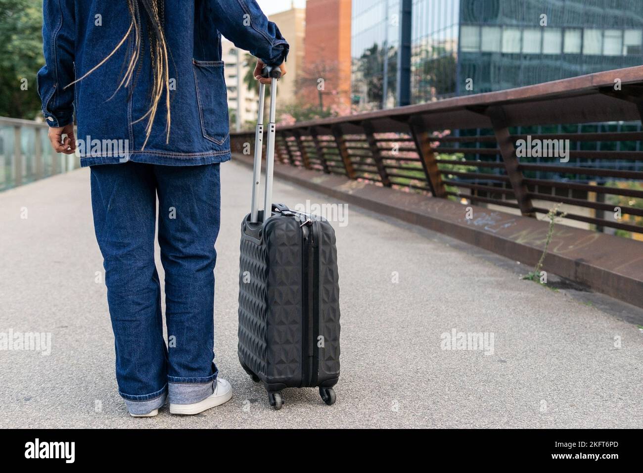 Back view of crop anonymous person in jeans and denim jacket with luggage standing on bridge near modern buildings Stock Photo